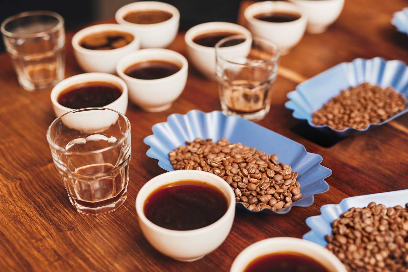 54726646 - many cups of coffee with open containers of freshly roasted coffee beans in a variety of flavours, and some water glasses on a wooden table for a tasting