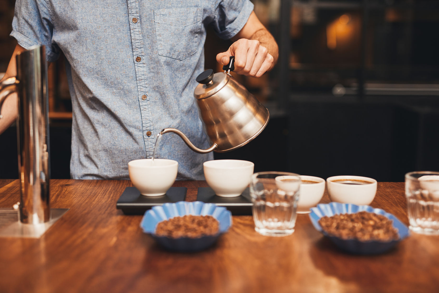 54726639 - professional barista pouring hot water into a cup of freshly ground coffee with open containers of coffee beans on a wooden counter in a roastery